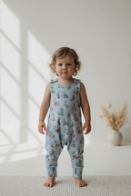 A toddler stands barefoot indoors wearing a light blue sleeveless one-piece outfit with an allover whimsical character print.snazzy sproglets