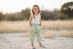 A young girl stands outdoors in a sunny field, wearing a sleeveless cream top and green floral pants with a white elastic waistband.