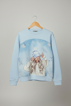 A light blue crewneck sweatshirt hanging on a wooden hanger against a neutral gray background. The sweatshirt features a large graphic print on the front depicting a girl in a winter outfit under a snowy arch. Snazzy Sproglets
