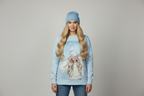 A woman modeling a light blue crewneck sweatshirt with a large graphic print of a blonde woman in a beanie, paired with a matching light blue beanie and blue jeans, against a solid gray studio background. Snazzy Sproglets