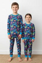 A boy and a girl stand barefoot indoors wearing matching long-sleeve pajama sets with a colorful dinosaur print on a blue background. The pajamas have bright blue trim at the collar, cuffs, and ankles.