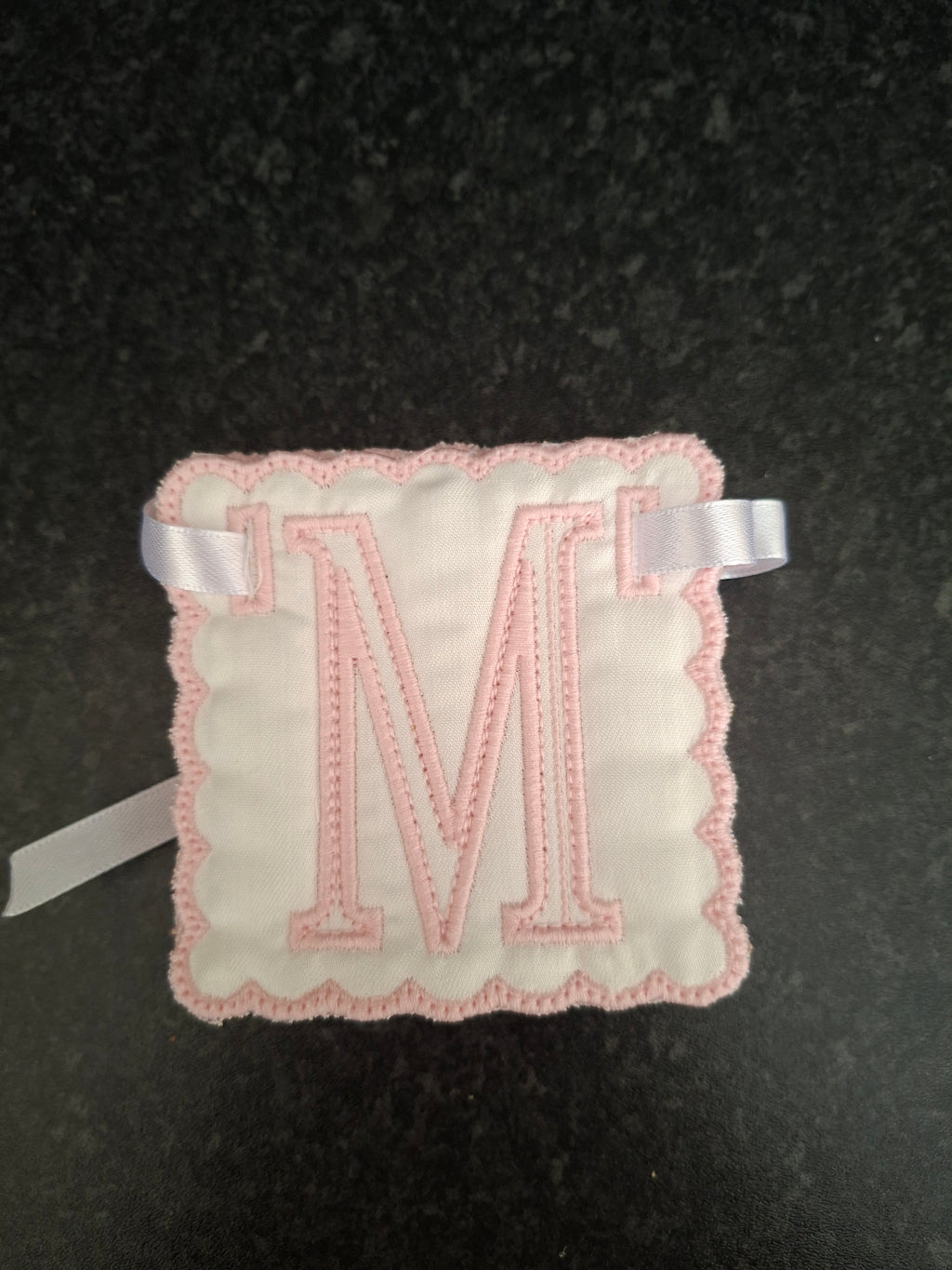 A square white fabric monogram patch with a scalloped pink border and a large pink embroidered letter 'M' in the center, finished with a white ribbon tab on each side snazzy sproglets