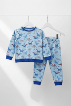 A two-piece children's pajama set hanging on white hangers against a light gray wall. The set includes a long-sleeve top and matching pants in light blue with a colorful dinosaur print and solid blue ribbed cuffs, collar, and hem. Snazzy Sproglets