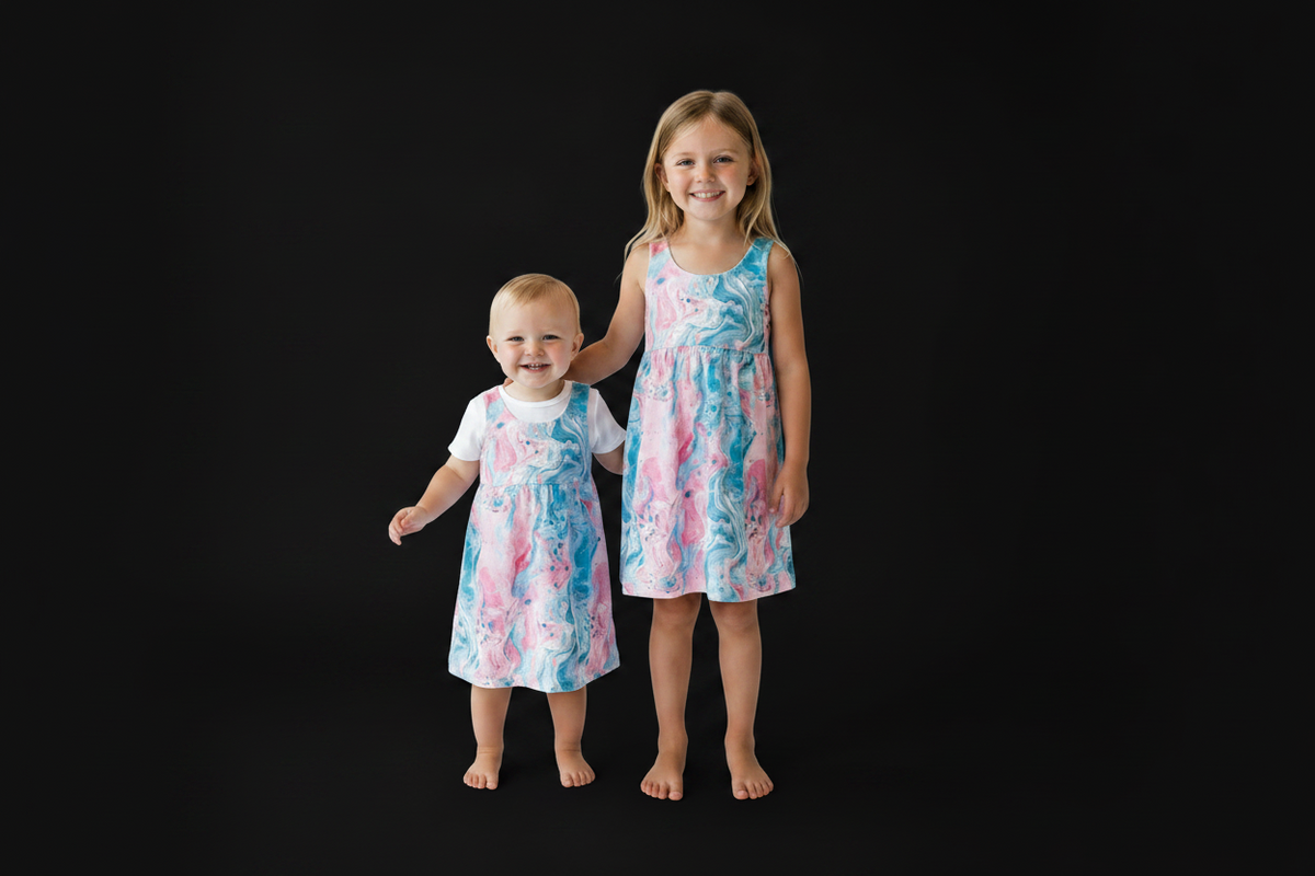 Two young girls standing barefoot against a solid black studio background. The older girl wears a sleeveless dress with a pink, blue, and white abstract watercolor pattern, and the younger girl wears a matching patterned dress over a white short-sleeve top.