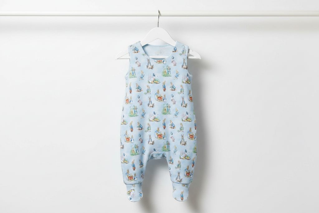 A light blue baby romper with an all-over Peter Rabbit print, hanging on a white hanger against a plain white wall. Snazzy Sproglets