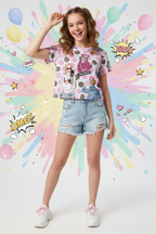 A short-sleeve graphic t-shirt with a pink, white, and gray polka dot pattern, featuring cartoon illustrations of women taking selfies, hearts, and text like 'I'm Cute' and 'Selfie'. The shirt is laid flat on a light wood background with green leaves in the top right corner.