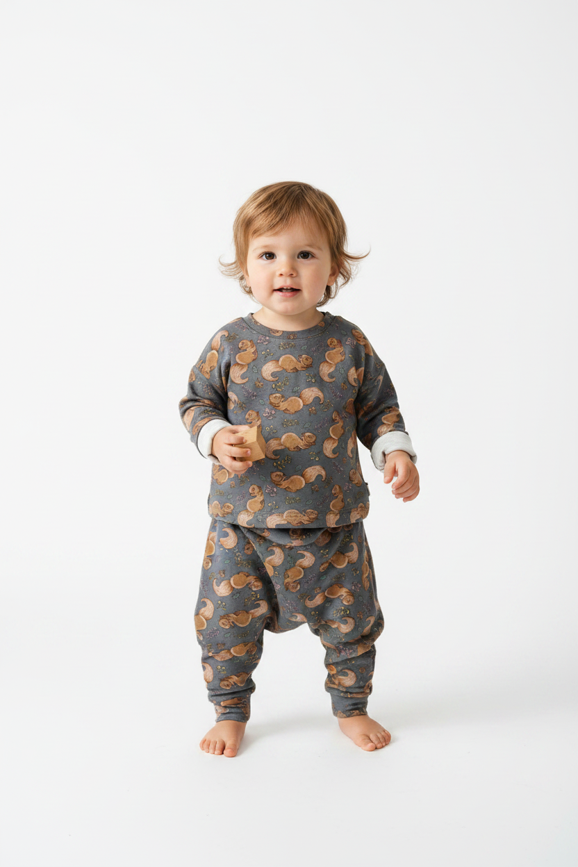 A toddler standing barefoot against a clean white studio background, wearing a matching long-sleeve top and pants set in dark gray with an allover brown squirrel print and light cuffs. Snazzy Sproglets