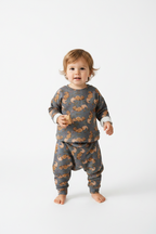 A toddler standing barefoot against a clean white studio background, wearing a matching long-sleeve top and pants set in dark gray with an allover brown squirrel print and light cuffs. Snazzy Sproglets