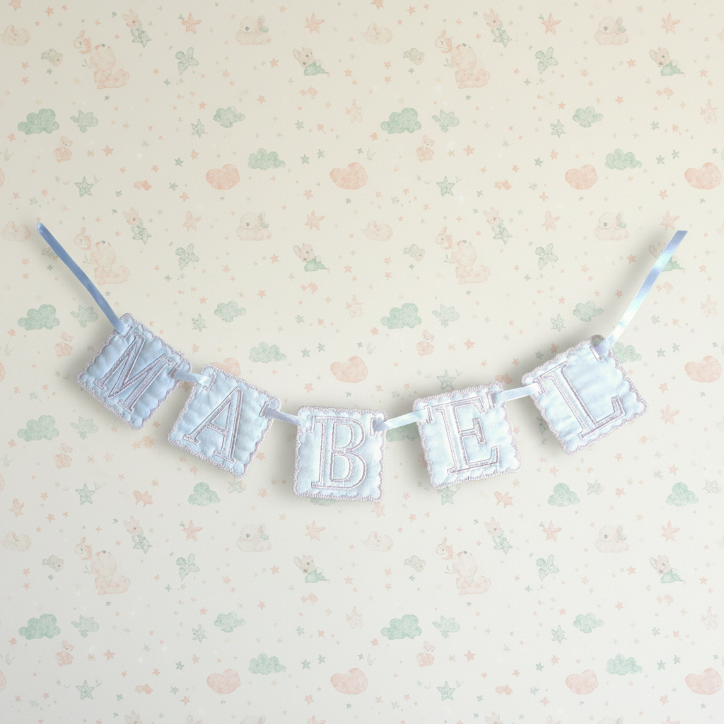 A fabric letter banner spelling 'MABEL' in light blue and white striped quilted letters, hung on a cream patterned wall.