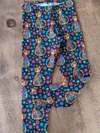 A pair of children's leggings with a colorful print of orange tabby cats, pink and blue flowers, and gold dots on a dark navy background, laid flat on a rustic wooden surface.