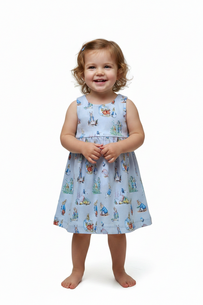 A young toddler girl stands barefoot against a plain white studio background, wearing a sleeveless light blue dress with a repeating Peter Rabbit print. snazzy sproglets