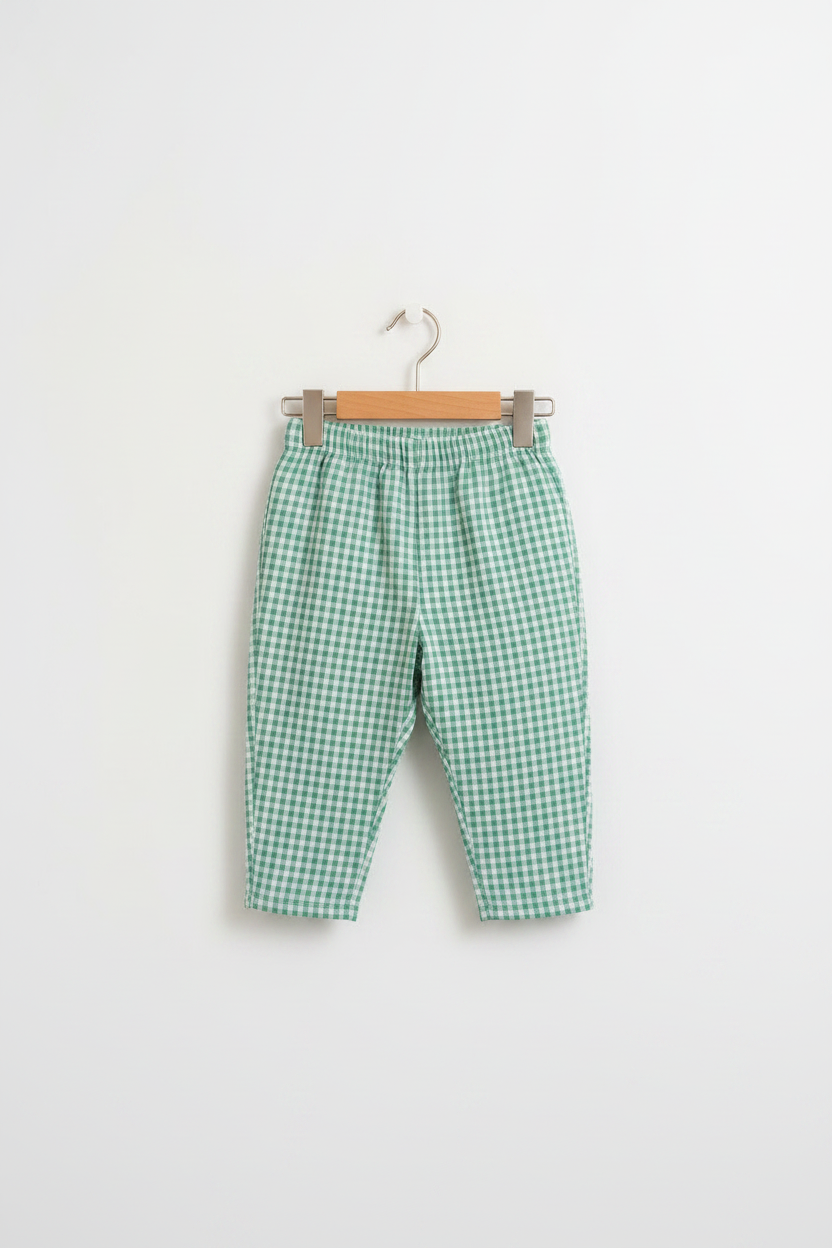 A young child with pigtails stands barefoot in a bright, modern room, wearing a loose white t-shirt and green-and-white gingham cropped pants.