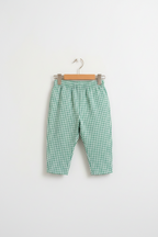 A young child with pigtails stands barefoot in a bright, modern room, wearing a loose white t-shirt and green-and-white gingham cropped pants.
