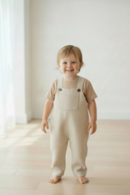 A smiling toddler standing barefoot on a light wood floor in a bright room, wearing a beige linen overall-style romper over a matching beige short-sleeve top. Snazzy Sproglets