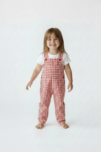 A young child standing barefoot against a plain white studio background, wearing red-and-white gingham bib overalls over a white short-sleeve top. Snazzy Sproglets