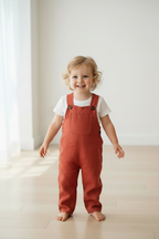 A smiling toddler standing barefoot on a light wood floor in a bright, minimalist room, wearing rust-orange linen overall over a white t-shirt. Snazzy Sproglets