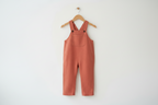 A terracotta-colored sleeveless baby jumpsuit or overall hanging from a wooden hanger against a plain white wall. Snazzy Sproglets