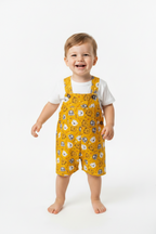 A smiling toddler standing barefoot against a clean white studio background, wearing mustard yellow overall-style shorts with a white T-shirt underneath and an allover smiley-face print. Snazzy Sproglets