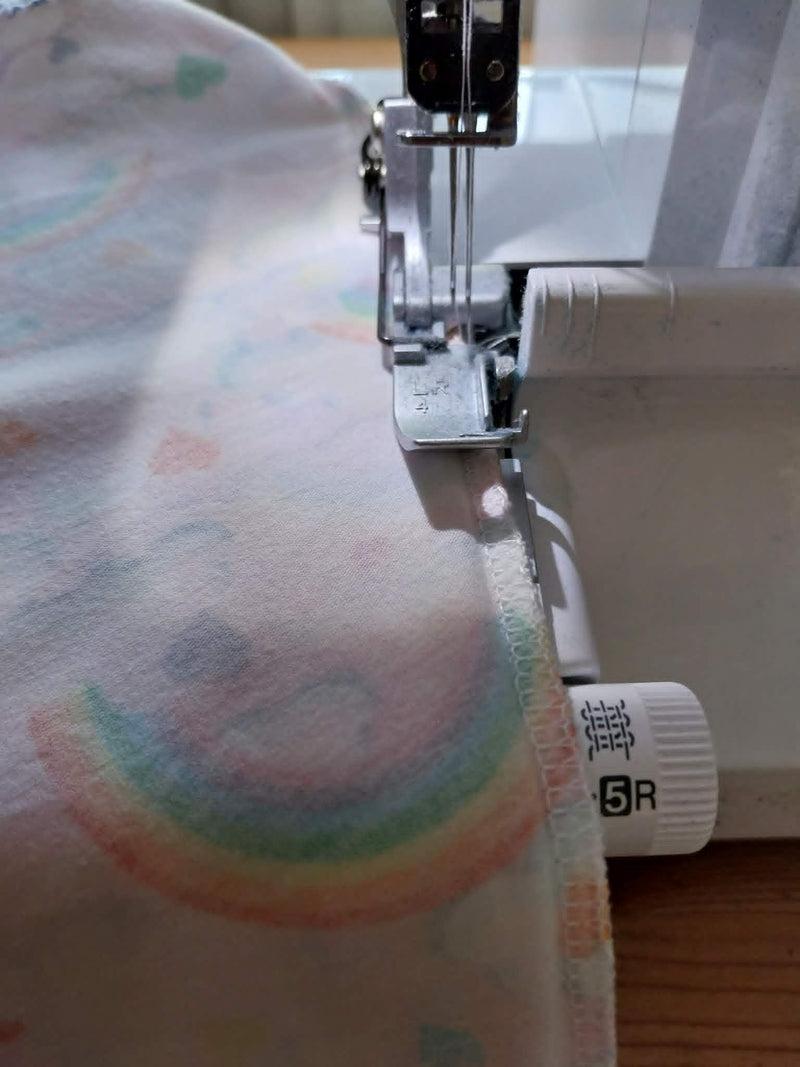 Close-up of a sewing machine stitching a soft fabric with a pastel rainbow print. The white machine body and metal press foot are visible, with a small white adjustment knob showing the brand name 'SR'.