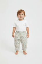 A toddler standing against a plain light gray studio background, wearing a plain white short-sleeve T-shirt and loose light-colored pants with a small floral print.