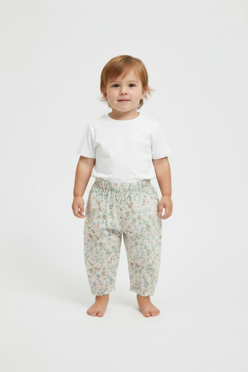 A toddler standing against a plain light gray studio background, wearing a plain white short-sleeve T-shirt and loose light-colored pants with a small floral print.