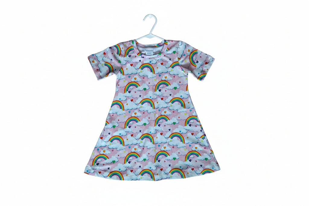 A short-sleeve baby t-shirt with a colorful repeating pattern of rainbows, clouds, and small hearts on a light pink background.