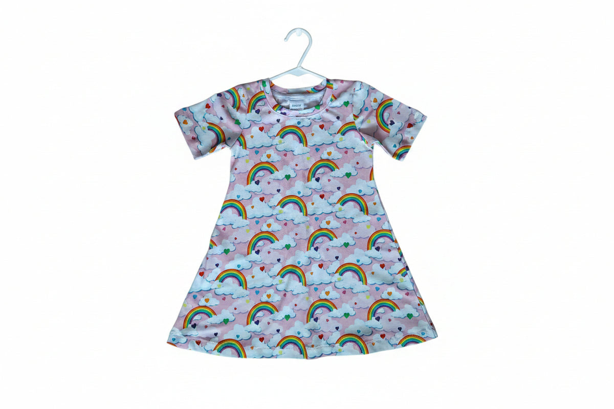 A short-sleeve baby t-shirt with a colorful repeating pattern of rainbows, clouds, and small hearts on a light pink background.