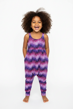 A young child with curly brown hair wearing a sleeveless zip-front romper with a purple, pink, and blue wavy pattern, standing against a plain white background. snazzy sproglets