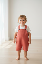 A smiling toddler stands barefoot on a light wood floor in a bright, minimalist room, wearing terracotta pink linen overall shorts over a white t-shirt with a sheep graphic. Snazzy Sproglets
