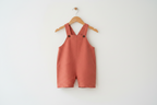 A terracotta-colored linen baby romper with an embroidered mouse on the chest, hanging from a wooden hanger against a plain white wall. Snazzy Sproglets