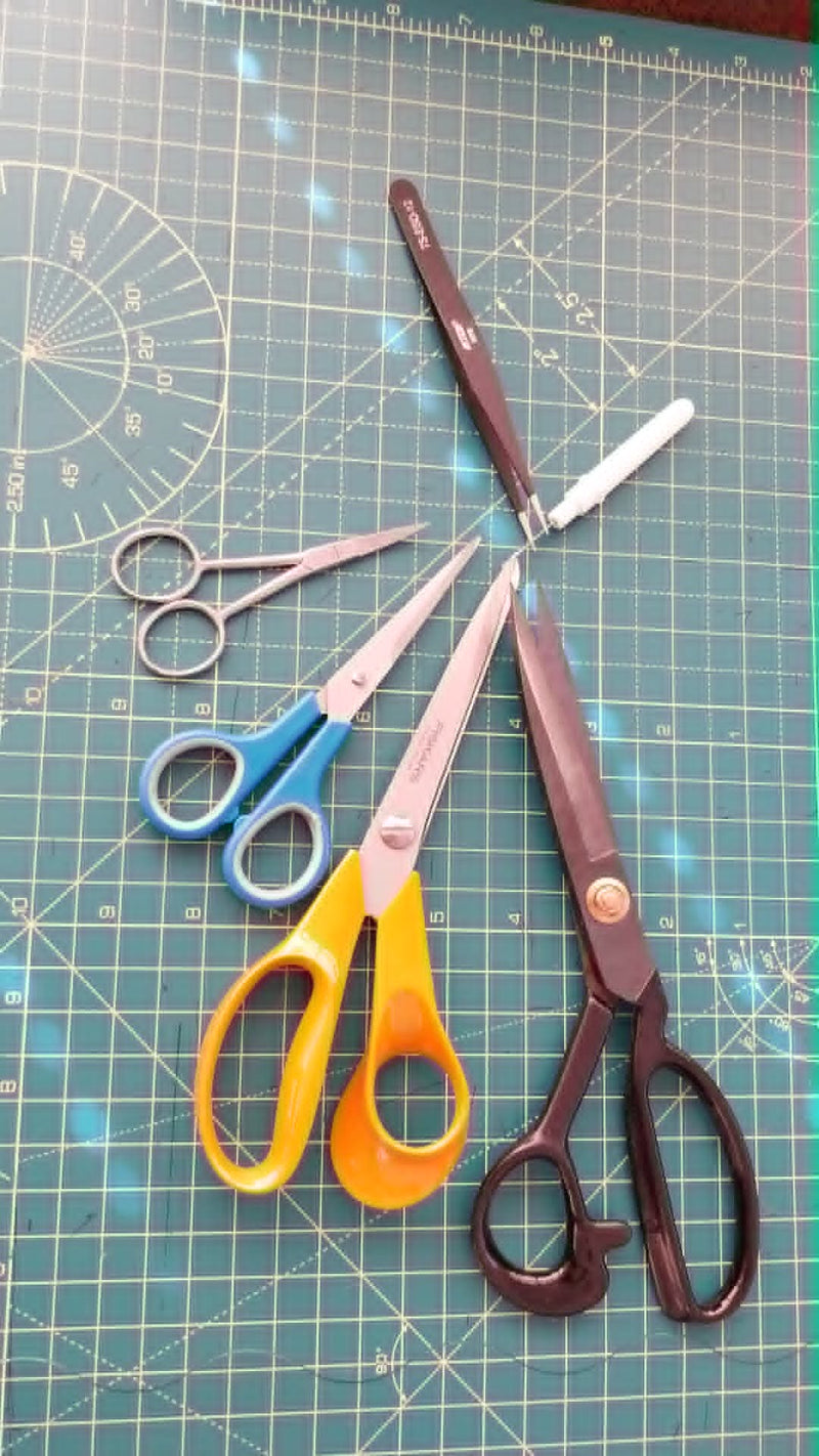 A collection of sewing and cutting tools arranged on a green gridded cutting mat, including large dark brown scissors, yellow-handled scissors, blue-handled scissors, silver ring shears, and a white-handled tool.