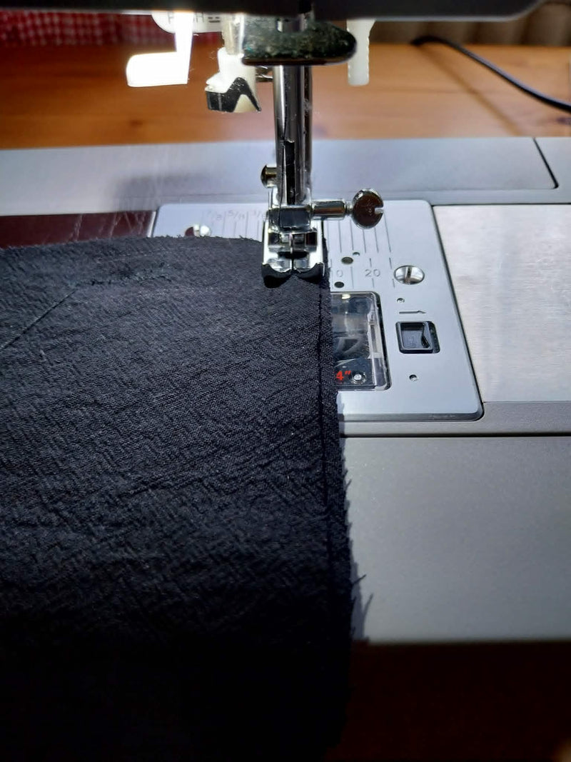 A close-up of a sewing machine in action, stitching a piece of dark charcoal gray textured fabric under the presser foot.
