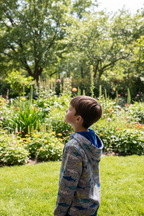 A young boy wearing a gray and blue patterned hoodie stands in profile in a lush green garden with flowers and trees in the background.