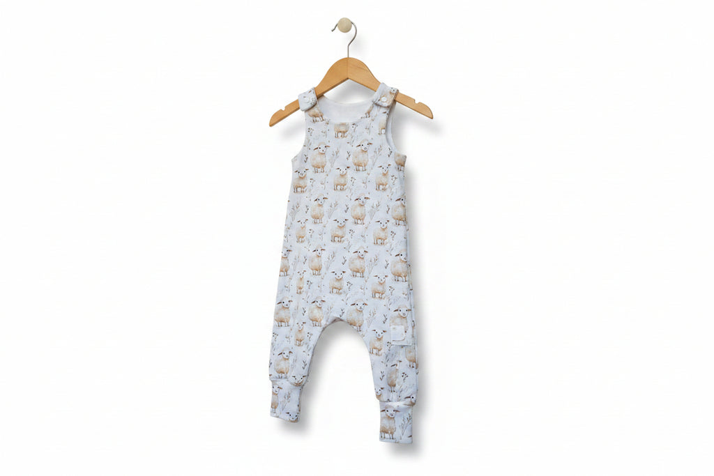A sleeveless baby jumpsuit with an allover sheep and wildflower print on a light blue-gray background, featuring adjustable shoulder straps and cuffed legs. snazzy sproglets