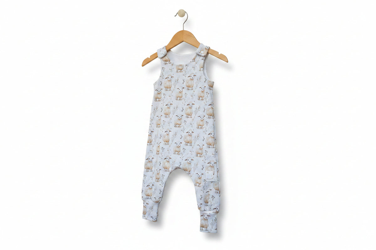 A sleeveless baby jumpsuit with an allover sheep and wildflower print on a light blue-gray background, featuring adjustable shoulder straps and cuffed legs. snazzy sproglets
