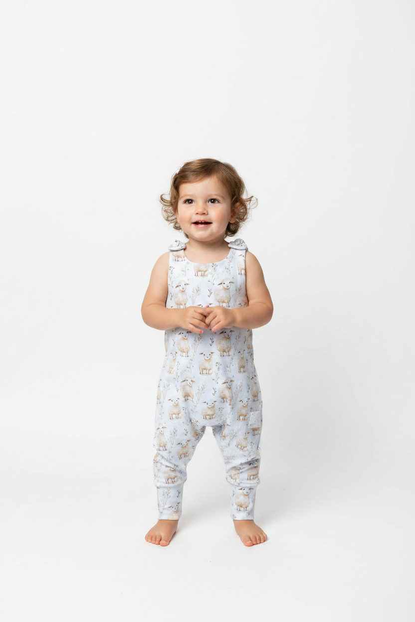A toddler girl stands barefoot against a seamless white studio background, wearing a sleeveless light blue overall romper covered in a small beige and brown sheep print. snazzy sproglets