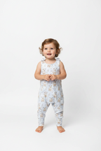 A toddler girl stands barefoot against a seamless white studio background, wearing a sleeveless light blue overall romper covered in a small beige and brown sheep print. snazzy sproglets