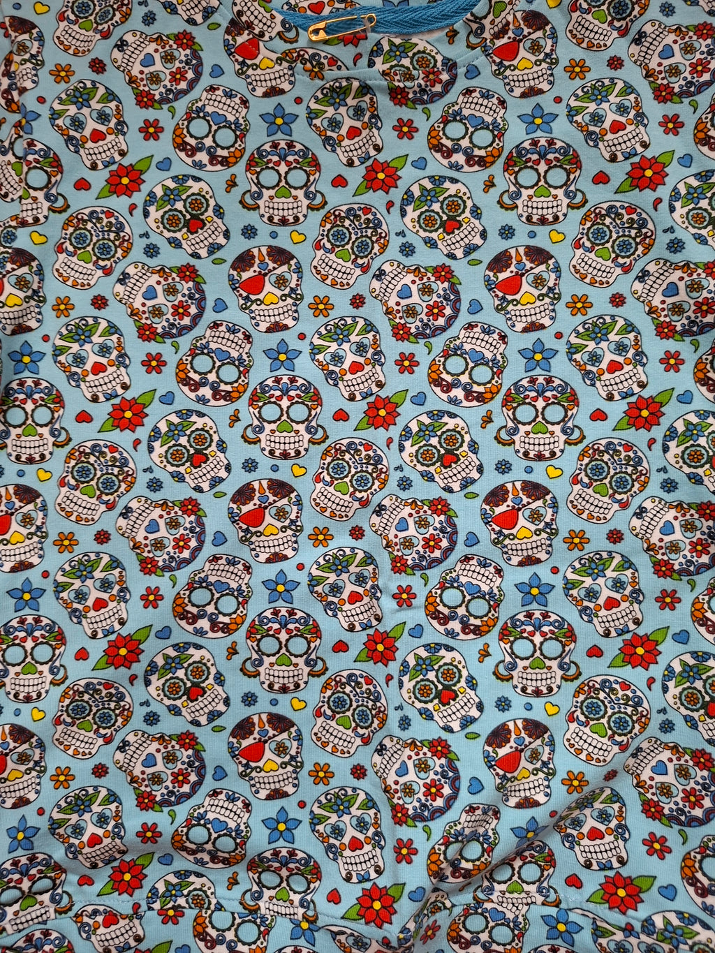 A close-up of a light blue fabric printed with a repeating sugar skull pattern in red, green, white, black, and blue, with small floral accents.