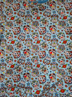 A close-up of a light blue fabric printed with a repeating sugar skull pattern in red, green, white, black, and blue, with small floral accents.