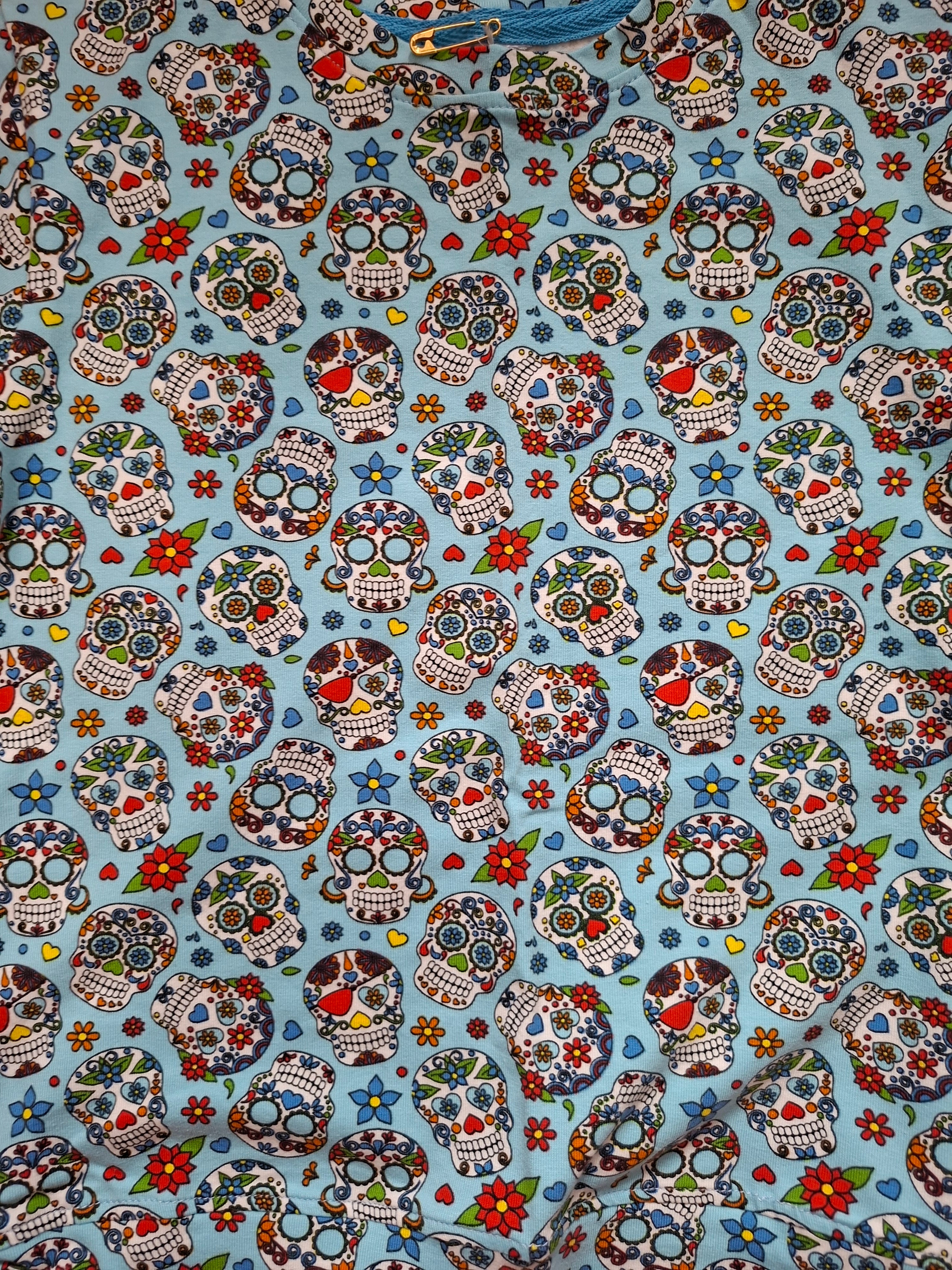 A close-up of a light blue fabric printed with a repeating sugar skull pattern in red, green, white, black, and blue, with small floral accents.