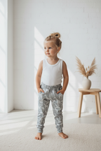 A toddler stands barefoot in a bright, sunlit room wearing a white sleeveless tank top and light gray patterned jogger pants with an elastic waist and cuffs.