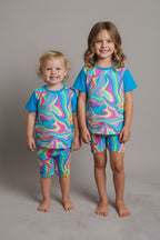 A two-piece kids'  set laid flat on a gray carpet. The set includes a short-sleeve top and matching pants with a bright blue collar and sleeves, and a body covered in a swirling psychedelic swirl pattern of pink, blue, yellow, green, and purple.