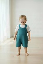 A smiling toddler standing barefoot on a light wood floor in a bright, minimalist room, wearing teal blue denim-style overall shorts over a white t-shirt. Snazzy Sproglets
