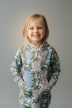 A young girl with blonde hair smiling, wearing a pastel rainbow hoodie with a green dinosaur and unicorn pattern, set against a dark gray studio background.