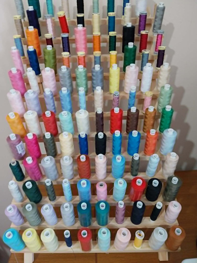A wooden thread display rack filled with dozens of colorful spools of thread, arranged in rows against a plain indoor wall.
